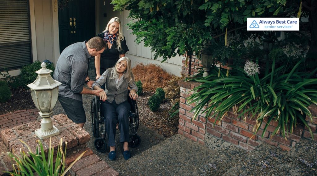 A caregiver and a family member are assisting an elderly woman in a wheelchair to safely navigate steps outside. They are outdoors, near a home with greenery and a lamp post in the background.