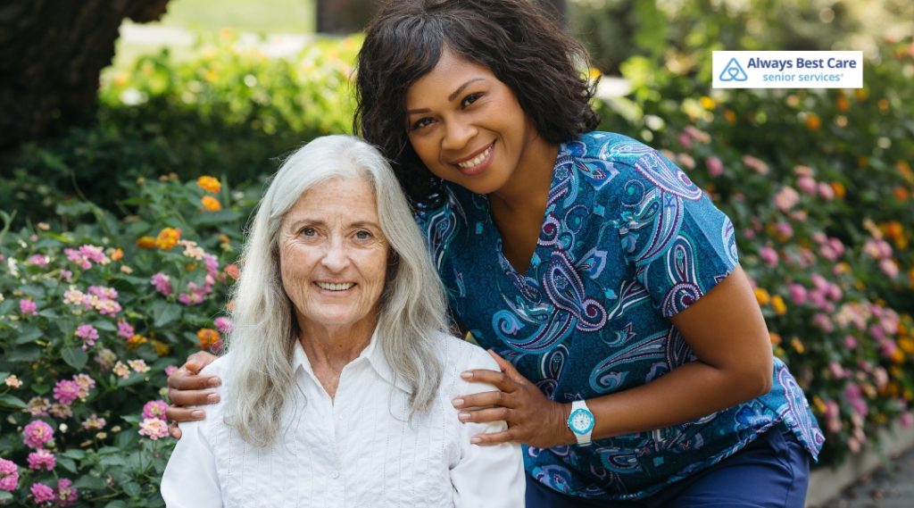 A caregiver and an elderly woman smile at the camera while standing outside among colorful flowers. The caregiver, wearing a patterned scrub shirt, has her arm around the elderly woman, who is sitting comfortably.