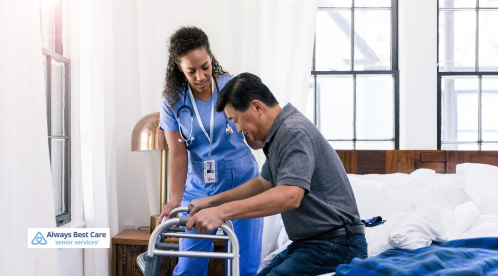 Female nurse assisting an older man as he carefully stands with a walker in a sunlit bedroom – Always Best Care senior services.
