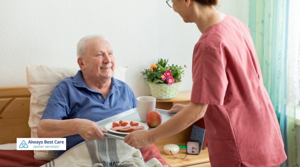 Smiling caregiver delivering a breakfast tray with fruit and toast to a senior man relaxing in bed – Always Best Care senior services.