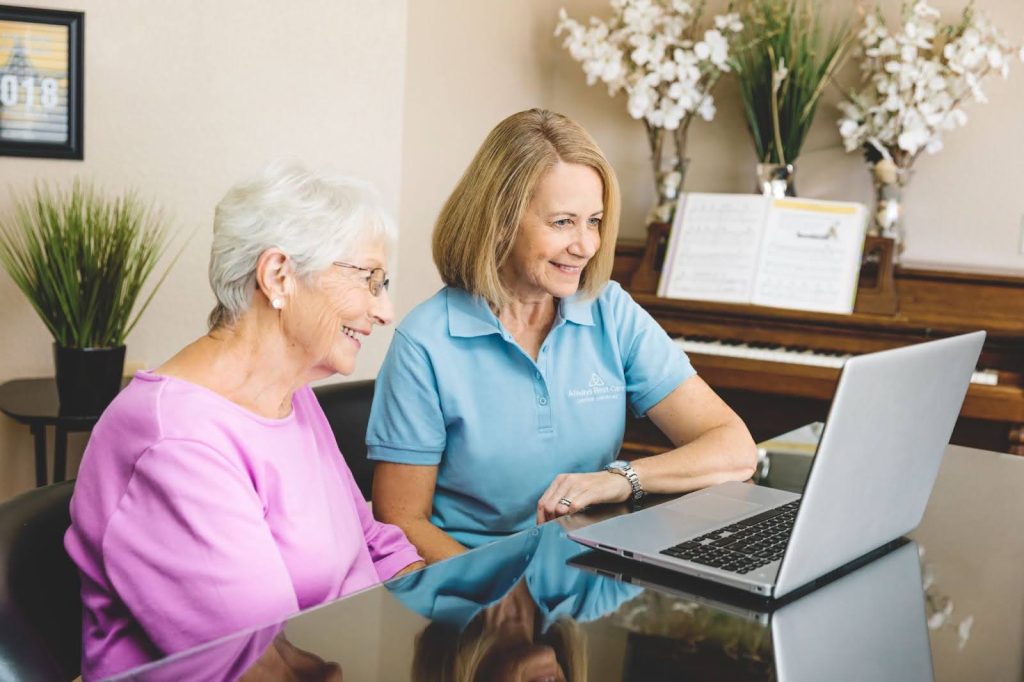 An older woman in a pink top sits beside a caregiver in a light blue Always Best Care polo shirt. They are smiling while looking at a laptop screen in a bright, homey room decorated with white flowers and a piano in the background.