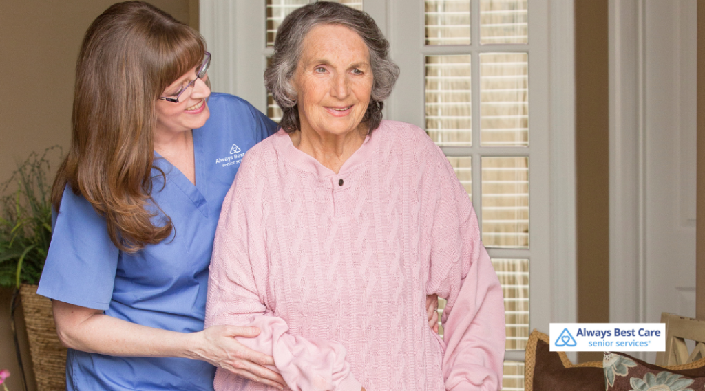 A caregiver with brown hair, wearing a blue uniform, assists an elderly woman with grey hair, dressed in a light pink sweater, as they walk together in a home setting. The Always Best Care logo is visible in the corner.