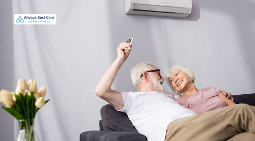 An elderly couple sitting together on a grey couch. The man wears glasses and holds up a remote control toward a wall-mounted air conditioning unit while smiling at his wife. Includes the "Always Best Care Senior Services" logo in the top-left corner.