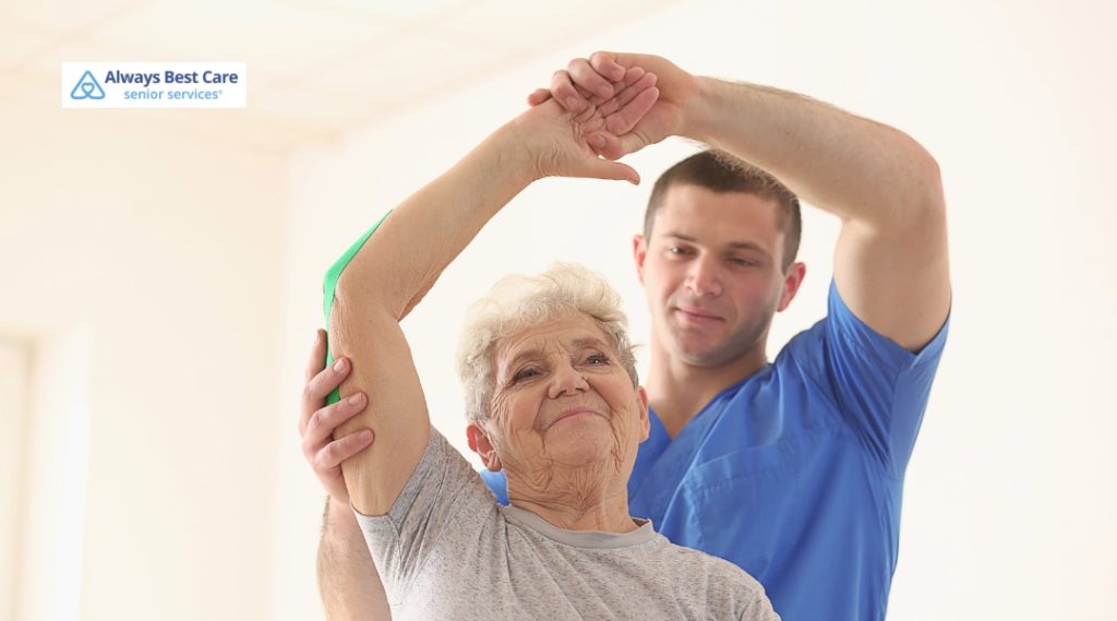 A caregiver assists a smiling elderly woman with gentle arm exercises for mobility and strength.
