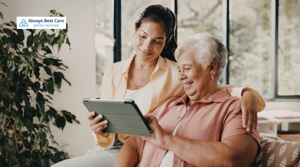 An older woman with short gray hair sits on a couch beside a younger woman with dark hair tied back. They are both smiling warmly while looking at a tablet that the older woman is holding. The younger woman has her arm gently resting around the older woman’s shoulder. An Always Best Care Senior Services logo is in the top left corner.
