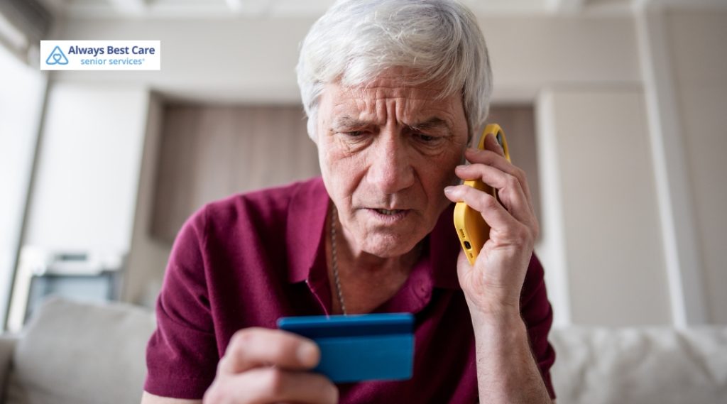 A close-up shot of an older man with white hair, looking stressed and confused as he holds a yellow smartphone to his ear. In his other hand, he holds up a blue credit card, staring down at it with a worried expression. An Always Best Care Senior Services logo is in the top left corner.