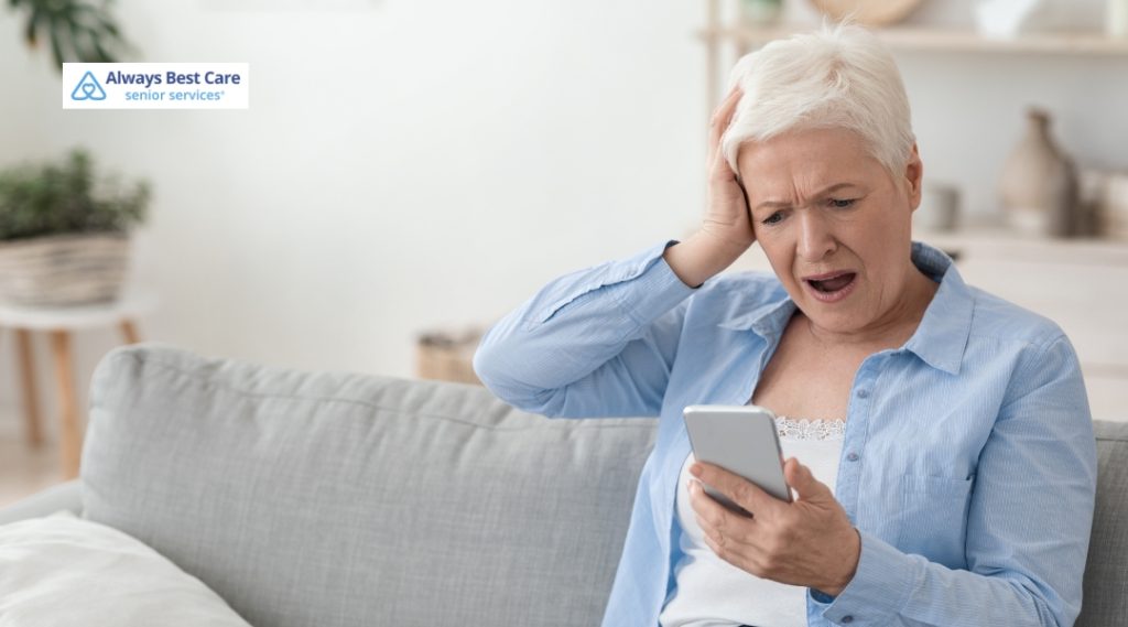 An older woman with short white hair sits on a gray couch, holding a smartphone in one hand with a shocked and distressed expression. Her other hand is raised to her head in frustration or disbelief as she looks at the phone screen. An Always Best Care Senior Services logo is in the top left corner.