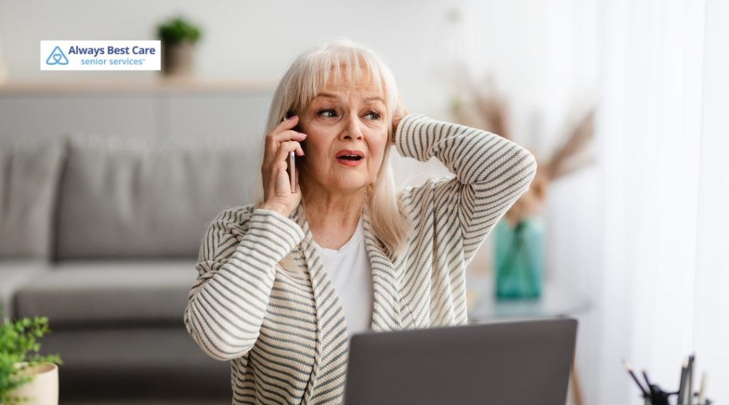 An older woman with blonde hair sits at a desk with an open laptop in front of her. She is holding a smartphone to her ear with a deeply concerned and panicked expression, clutching the back of her head with her other hand. An Always Best Care Senior Services logo is in the top left corner.
