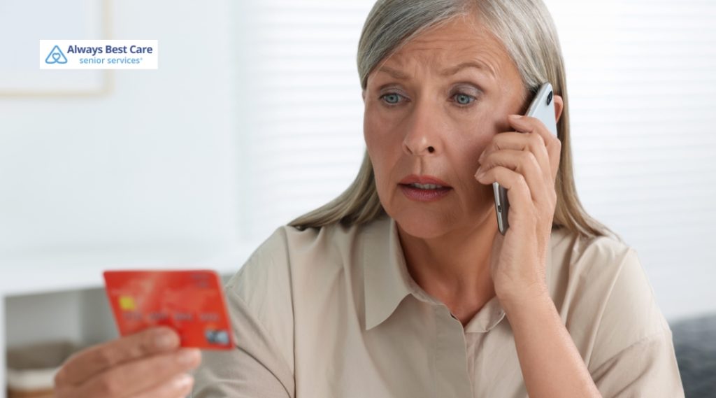A close-up shot of an older woman with gray hair looking anxious and uncertain while talking on a silver smartphone. She is holding a red credit card up in her other hand, looking toward it with concern. An Always Best Care Senior Services logo is in the top left corner.v