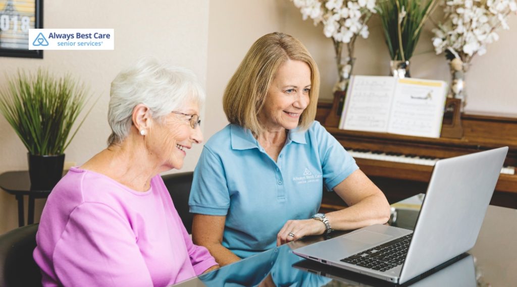 An older woman in a pink shirt and a female caregiver wearing a light blue Always Best Care polo shirt sit side-by-side at a shiny dark table. They are both smiling as they look at an open laptop screen together. A piano is visible in the background.