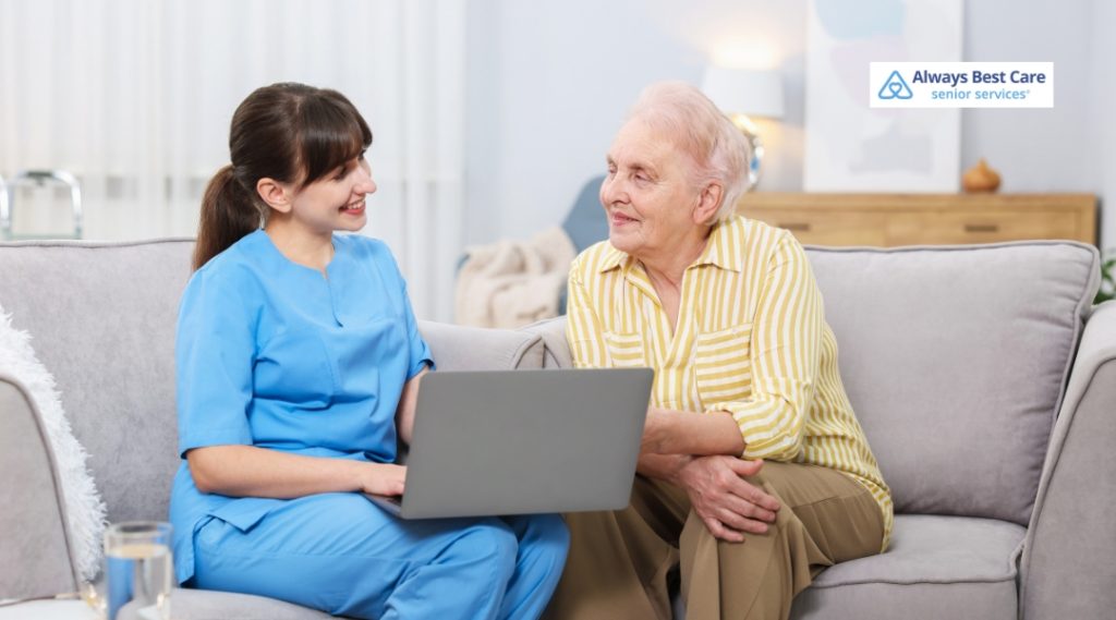 A female caregiver in blue medical scrubs sits on a gray sofa next to an older woman in a yellow striped shirt. The caregiver is smiling and looking at the older woman while holding a laptop on her lap. The older woman smiles back comfortably. An Always Best Care Senior Services logo is in the top right corner.