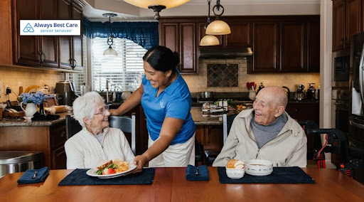 In a warm kitchen setting, a smiling caregiver in a blue polo shirt serves a plate of food to an elderly woman and an elderly man seated at a wooden dining table. Both seniors are smiling back at her. The Always Best Care Senior Services logo is visible in the top left corner.
