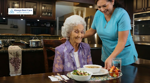 A caregiver in a blue uniform stands beside a seated elderly woman with white hair, adjusting her meal of soup, salad, and fresh fruit on a dining table. Both are smiling cheerfully in a home environment. The Always Best Care Senior Services logo is in the top left corner.
