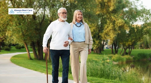 An elderly man with a beard and a cane walking arm-in-arm with an elderly woman along a paved park path next to a pond. Includes the "Always Best Care Senior Services" logo in the top-left corner.