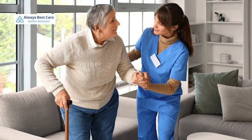 A female caregiver in blue scrubs holding the hand of an elderly woman as she stands up from a grey couch using a cane. Includes the "Always Best Care Senior Services" logo in the top-left corner.
