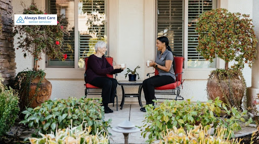 An elderly woman and a female caregiver sitting in red chairs on a house patio, drinking from mugs and talking. Potted plants and bushes decorate the patio space. Includes the "Always Best Care Senior Services" logo in the top-left corner.