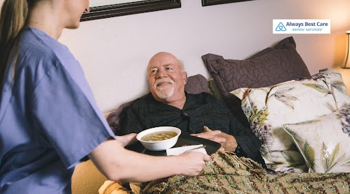 A caregiver in blue scrubs brings a meal tray containing a bowl of warm soup to a smiling, white-bearded senior man propped up with pillows in bed. The "Always Best Care Senior Services" logo is positioned in the top-right corner.