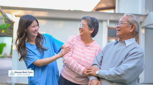 A young female caregiver in blue scrubs smiling and walking outside with an elderly East Asian couple who are holding hands and laughing. Includes the "Always Best Care Senior Services" logo in the bottom-left corner.
