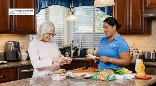 A caregiver in a blue polo shirt spreads condiments on bread while a senior woman with blonde hair and glasses prepares sliced lunch meat beside her at a kitchen island. Ingredients like lettuce, tomatoes, and bread line the counter. The "Always Best Care Senior Services" logo is in the top-left corner.
