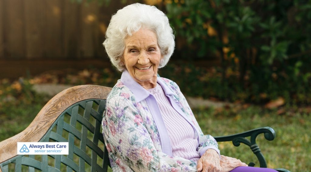 An elderly woman sitting on a park bench, smiling and enjoying the outdoors. She looks content, with natural sunlight and a lush green background enhancing the peaceful mood.
