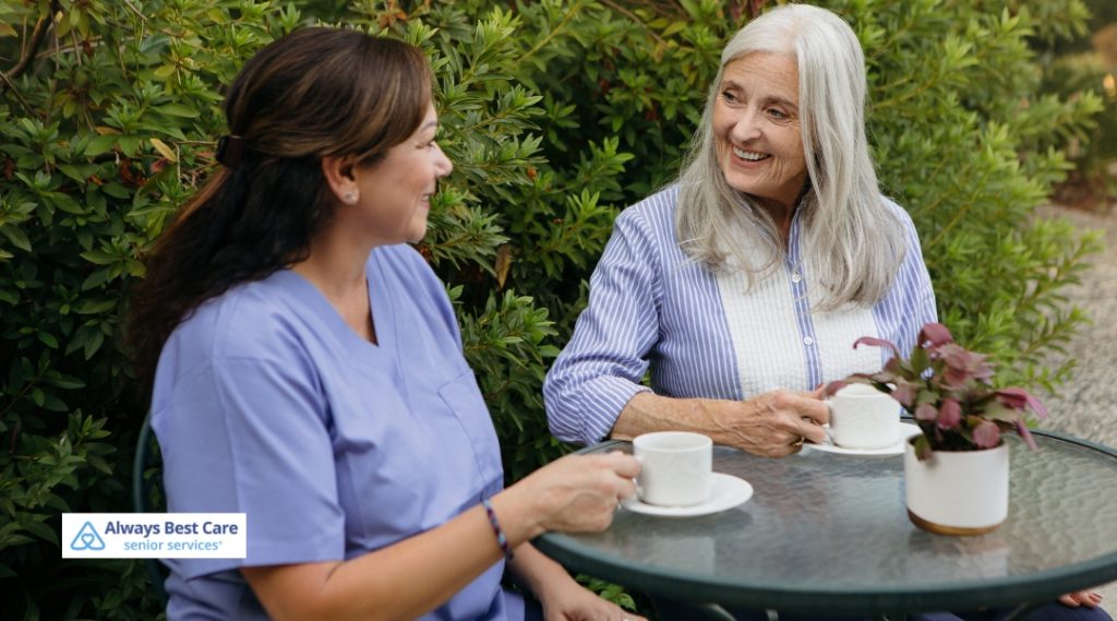 Two women, one an elderly individual and the other a caregiver, enjoying a conversation over coffee at a garden table. The scene conveys warmth, relaxation, and companionship in an outdoor setting.