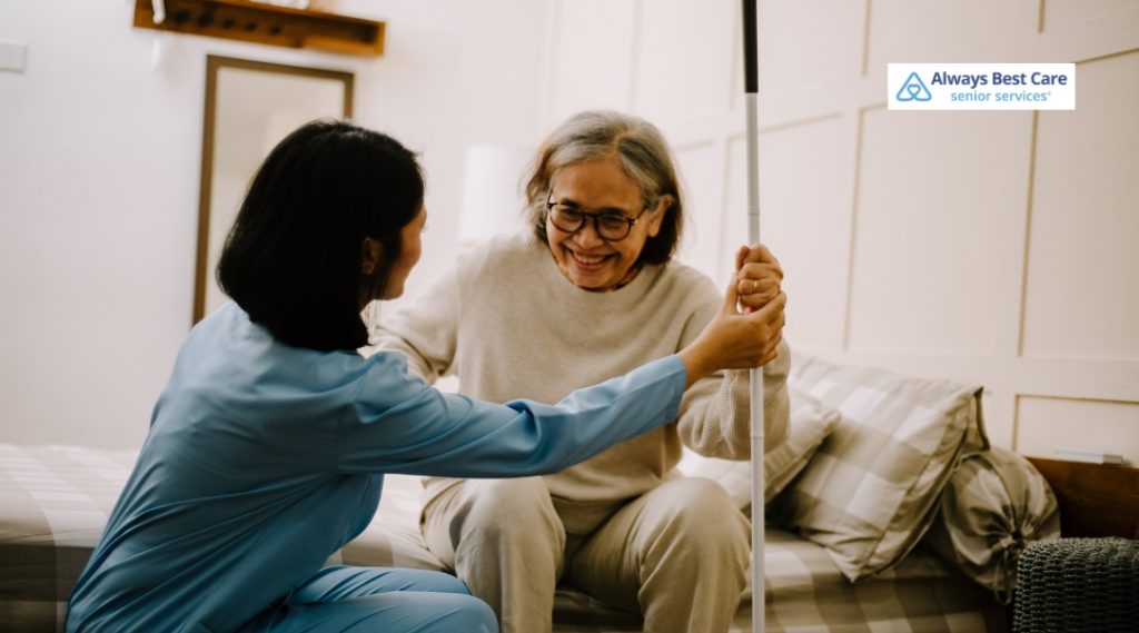 Smiling senior woman sitting on a bed assisted by a caregiver with a walking cane, capturing supportive mobility care in a cozy home setting.