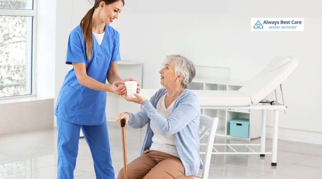 Young caregiver handing a cup to an elderly woman with a cane in a bright medical room, demonstrating helpful daily support and hydration care.