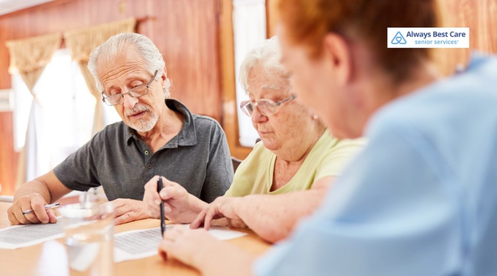 Senior couple reviewing documents with a caregiver at a table, illustrating family involvement and planning in senior care decisions.