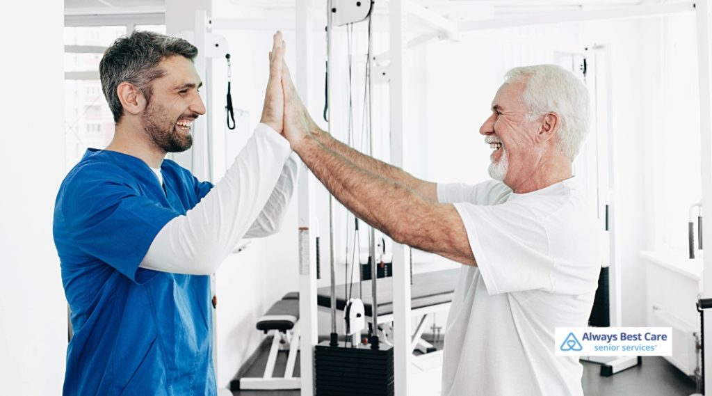 A caregiver and elderly man share a high-five and smile in a rehabilitation or exercise setting.