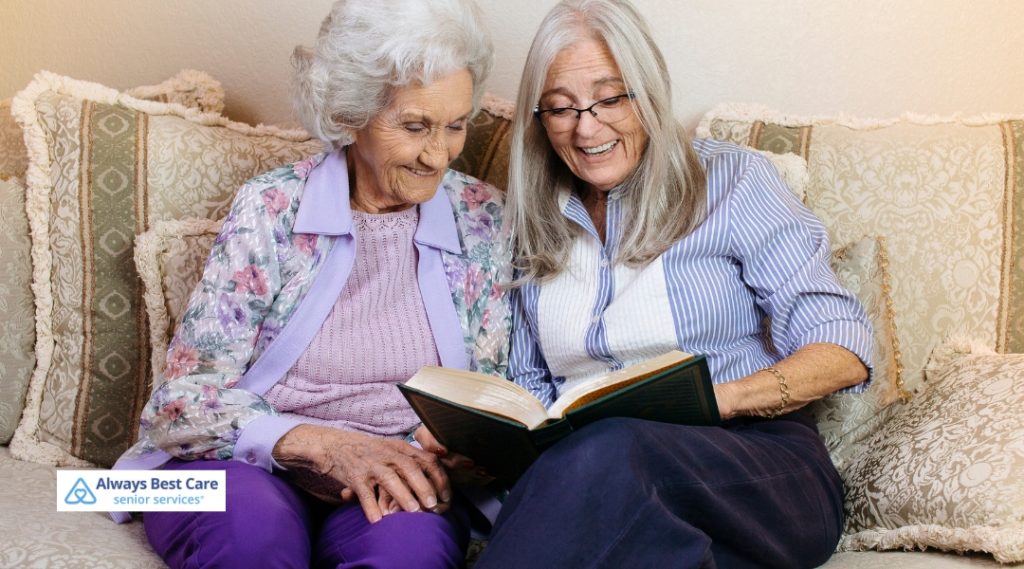 An elderly woman and her caregiver sitting on a cozy sofa, smiling and reading a book together. The scene is warm, showing a moment of connection and companionship.