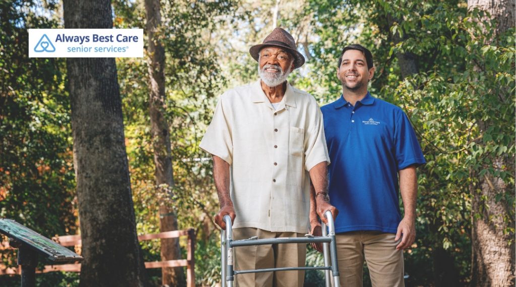 An elderly man with a walker and a male caregiver stand together on a wooded path, smiling and enjoying outdoor time as part of personalized senior care.