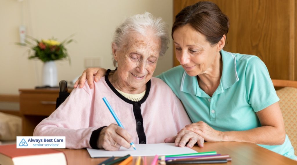 A senior woman is seated at a table, coloring with crayons as a caregiver gently watches her. The caregiver is offering support and guidance, and the woman looks happy and engaged in the activity. The setting is cozy and warm, with books and art supplies around them.
