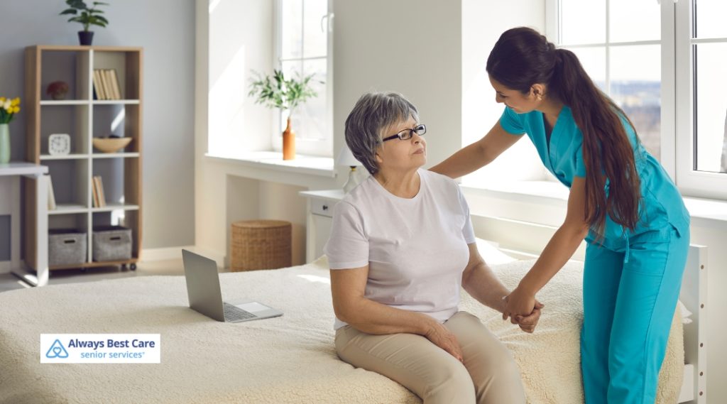 A caregiver is helping a senior woman with mobility support, gently guiding her as she stands from a seated position in a bright and modern bedroom. The caregiver is wearing scrubs and the elderly woman is dressed in a casual white shirt, showing a caring and supportive moment.