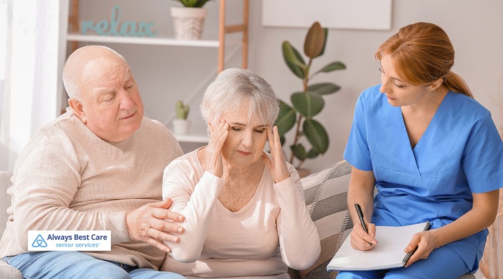 A couple is sitting on a couch, with the woman holding her head, seemingly distressed. The man beside her is offering comforting support, while a caregiver, taking notes, is observing the situation. The scene conveys care and attention to the elderly woman’s well-being.