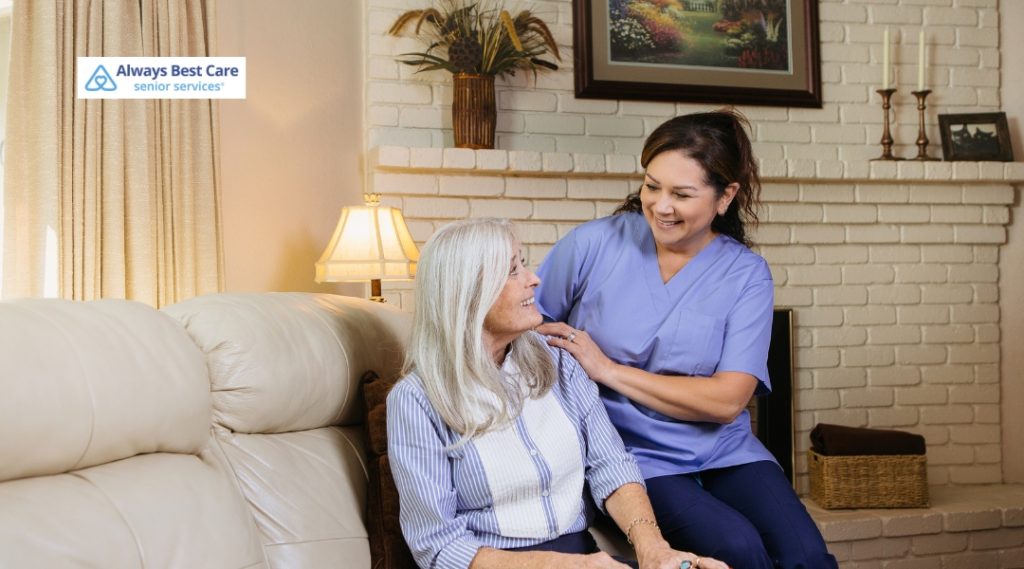 A senior woman and her caregiver, smiling and engaging in a pleasant conversation while sitting on a couch in a cozy, well-decorated living room. The Always Best Care logo is in the top left corner