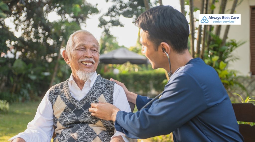 Caregiver checking an elderly man’s heart with a stethoscope outside, both smiling warmly, highlighting outdoor wellness care and trust.