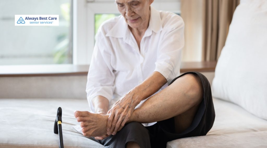 An elderly woman in a white shirt sits on a couch, holding her foot while assessing discomfort, with a cane next to her.