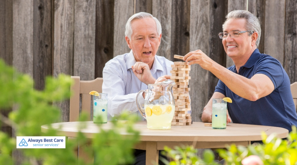 Two older men are playing a game of Jenga outside on a sunny day. Both men are smiling and engaged, with a pitcher of lemonade and drinks on the table, and the Always Best Care logo in the corner.