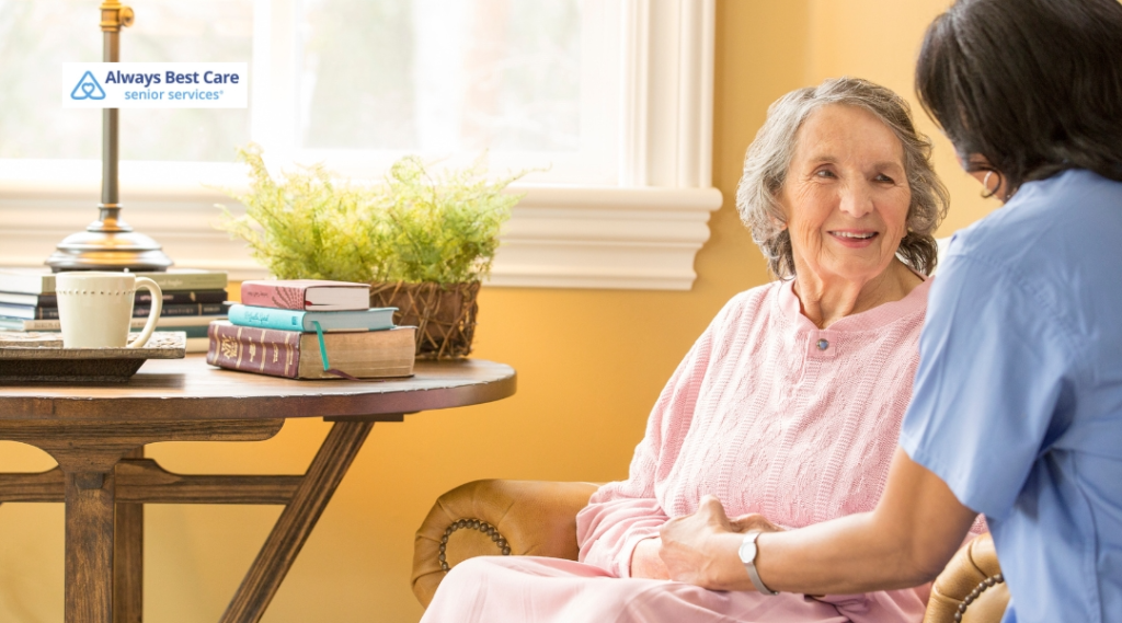 A senior woman sits comfortably on an armchair, smiling and talking with a caregiver in a cozy, well-lit room. Books and a cup of coffee are placed on a small table next to them, with the Always Best Care logo in the corner.