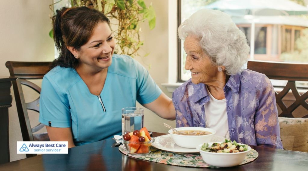 A caregiver and an elderly woman share a meal together at a dining table. The caregiver, dressed in a blue shirt, is smiling and engaging with the woman, who is enjoying a bowl of soup and salad. The Always Best Care logo is in the corner.