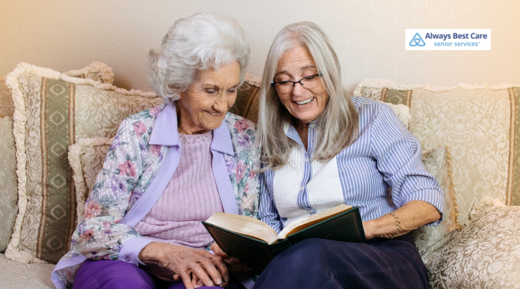 An elderly woman and a caregiver smile while looking at a book together, sitting on a couch with soft, cozy pillows around them. Both women appear joyful and engaged, with the Always Best Care logo visible in the corner.