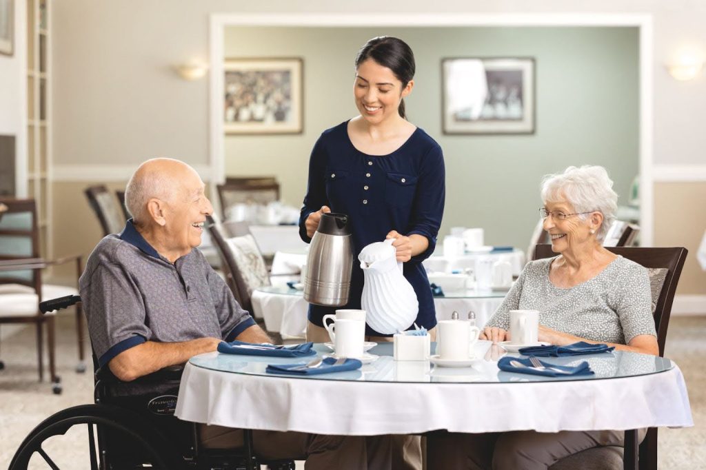 An older couple smiles at each other while seated at a dining table as a cheerful caregiver pours coffee for them inside a bright, welcoming dining room.